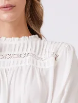 Blusa azul de gasa con pliegues en la parte delantera y detalles de encaje calado. Cuello alto fruncido y mangas largas con puños elastizados. Espalda lisa con abertura tipo gota y botón.