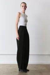 Pantalón negro de tiro alto, con pliegues en el delantero y bolsillos laterales ocultos. Cierre frontal con cremallera y ganchos metálicos.