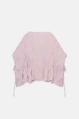 Top blanco de cuello redondo y mangas tipo capa, con textura fruncida, bajo asimétrico, lazos decorativos en el escote y en los laterales, y cierre posterior con botón.