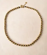 Collar de diseño minimalista compuesto por una secuencia de esferas doradas intercaladas con nudos de hilo negro. Presenta un cierre de mosquetón con cadena de alargue y un pequeño dije circular grabado.
