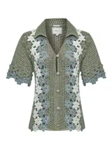 Sweater verde de tejido tipo crochet, con cuello camisero, mangas cortas y cierre frontal de botones. Presenta un diseño de bandas laterales con aplicaciones florales en tonos blancos y celestes.