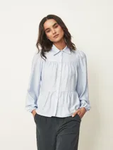 Camisa de corte amplio y estilo romántico, color celeste, con cuello clásico, cierre frontal de botones y diseño de canesú fruncido. Presenta mangas largas abullonadas con puños elásticos.