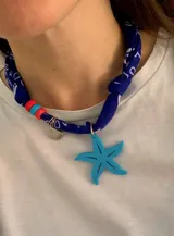 Collar tipo gargantilla confeccionado con un pañuelo bandana azul con estampado blanco. Presenta un dije central en forma de estrella de mar color turquesa, un dije metálico pequeño en forma de concha y cuentas de colores rojo y turquesa.