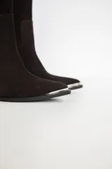 Bota bucanera de cuero color marrón, de caña alta, con taco cuadrado negro y punta fina con detalle metálico. Presenta tiras laterales con tachas y herraje metálico.