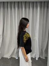 Remera negra de corte regular con estampado gráfico en color amarillo. Presenta inscripciones con estética oriental en la parte superior izquierda y en el lateral derecho. Confeccionada en algodón de alto gramaje con cuello reforzado y grifa de la marca en la manga.