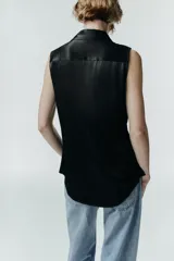 Camisa sin mangas de tejido satinado, con cuello solapa, cierre frontal con botones ocultos y bolsillos de plastrón con solapa en el delantero. Presenta un bajo asimétrico y corte recto.