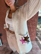 Saco tejido en lana color crudo, con corte recto, mangas largas y bolsillos frontales con apliques de estrellas de cuero en marrón, rosa y animal print.