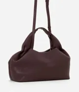 Cartera mediana de diseño estructurado en color bordó, confeccionada en poliuretano con acabado liso. Presenta asas de mano integradas con detalle fruncido, correa larga ajustable y desmontable, y cierre superior con cremallera.