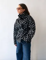 Campera de algodón con estampado de lunares blancos sobre fondo negro. Presenta cuello alto, cierre frontal con botones metálicos y cinturón ajustable en la cintura. Posee bolsillos laterales y un diseño versátil que permite llevarla entallada o con silueta holgada.