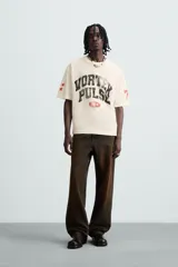 Camiseta de manga corta color crema, corte boxy fit, con cuello redondo. Presenta un estampado frontal en negro con el texto 'VORTEX PULSE' y el número '1974' en un óvalo rojo desgastado. Tiene pequeños detalles gráficos rojos en las mangas.