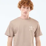 Camiseta de algodón color beige con cuello redondo y manga corta. Presenta un pequeño estampado frontal de una calavera con sombrero de paja y huesos cruzados.