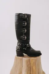 Bota alta de cuero negro con caña recta y puntera cuadrada. Presenta un taco bajo y múltiples correas con hebillas metálicas plateadas.