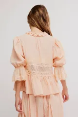 Camiseta de tela liviana color salmón, con terminaciones de encaje, escote en V profundo y botonadura al frente con tiras para anudar. Mangas tres cuartos abullonadas con puño elastizado y ruedo acampanado, cuello con volado.