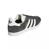 Championes adidas Gazelle de diseño clásico, confeccionados en gamuza color gris oscuro con las icónicas tres tiras laterales en blanco. Presentan suela de goma blanca, lengüeta blanca con logo y detalle de marca en dorado en el lateral.