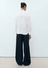 Camisa de lino blanca de corte recto con cuello camisero, manga larga con puños abotonados, cierre frontal de botones y un bolsillo de parche en el pecho.