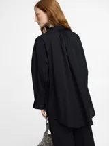 Camisa negra de corte oversize y manga larga con puños abotonados. Presenta un cuello clásico con un lazo fino para anudar en la parte superior y un diseño con bajo asimétrico, siendo más larga en la parte posterior.