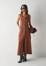 Vestido largo de sarga en color terracota, sin mangas, con escote redondo y cierre frontal completo de botones. Presenta una silueta entallada con bolsillos laterales y costuras verticales que definen el diseño.