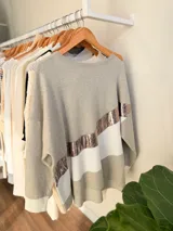 Buzo de tejido de punto con diseño oversize y calce relajado. Presenta un diseño de bloques de color en tonos neutros con una franja diagonal central en material metalizado brillante. Cuenta con cuello redondo y mangas largas con puños acanalados.
