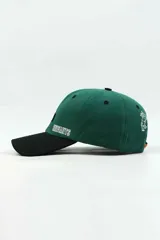 Gorra con visera de diseño bicolor, con cuerpo en color verde y visera en color negro. Presenta un parche rectangular con un punto azul en el centro y la palabra 'ENFANTS' bordada en blanco en el panel frontal.