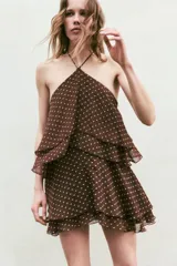 Vestido mini de cuello halter con diseño de volantes escalonados. Presenta estampado de lunares, cierre lateral con cremallera oculta y lazada ajustable en el cuello.