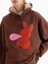 Buzo marrón de corte amplio con capucha forrada en corderito. Presenta una estampa frontal de un conejo rojo haciendo una burbuja de chicle, parte de la cápsula Il Bunny Porta Fortuna.