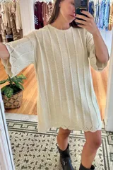 Vestido corto de tejido de punto color crema, con diseño de trenzas verticales, mangas cortas acampanadas y terminaciones con borde ondulado.