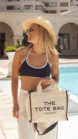 Bolso tipo tote bag de lona color beige con detalles de ribetes negros. Presenta el texto 'THE TOTE BAG' y 'LINCOLN'S' estampado en el frente. Cuenta con doble asa superior y correa ajustable extraíble.