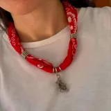 Collar tipo bandana de tela roja con estampado paisley blanco, adornado con anillos metálicos plateados y un dije colgante en forma de oso con incrustaciones de cristales.