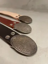 Cinto de cuero con hebilla ovalada de metal con diseño labrado. Disponible en color natural, marrón y bordó.