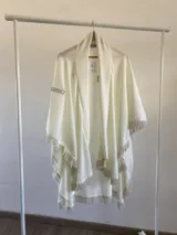 Ruana de tela fina con textura, color natural, con diseño abierto y terminaciones con flecos en todo el borde. Incluye una etiqueta decorativa en la parte superior.
