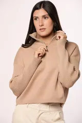 Buzo tejido de punto color beige, con cuello alto y cierre frontal parcial. Presenta mangas largas con diseño en tejido morley y corte holgado.