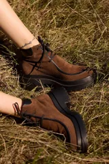 Botas de caña corta confeccionadas en cuero nobuk color marrón, con detalles en cuero negro en los bordes y cordones negros. Presentan una suela de plataforma dentada de color negro.