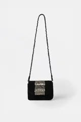 Cartera bandolera de cuero negro con solapa, decorada con apliques bordados de lentejuelas y abalorios plateados en el centro. Incluye una correa larga de hombro estilo cadena trenzada.