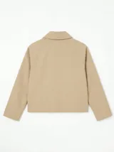 Chaqueta corta de gabardina en color beige, con cuello camisero, cierre frontal de botones y mangas largas con puños vueltos que revelan un forro a cuadros.