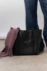 Bolso tipo tote de color negro con textura granulada, diseño amplio y funcional con asas largas para llevar al hombro. Presenta el logo de la marca en la parte frontal.