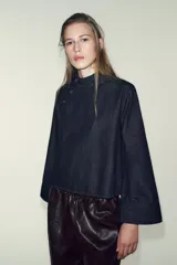 Camisa de denim azul oscuro, de corte cuadrado y manga ancha acampanada. Presenta cuello mao y cierre asimétrico con botones a presión en el frente.