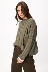 Sweater de punto con diseño de trenzas verticales, cuello redondo y corte crop. Presenta un parche decorativo en el lateral y una textura tejida con relieve.