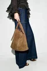 Bolso tipo saca de diseño asimétrico confeccionado en piel de ante. Cuenta con un asa bandolera ajustable, cierre mediante imán y un bolsillo interior con cremallera.