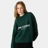 Suéter de punto grueso color verde oscuro, con corte relajado y ligeramente corto. Presenta el logo de la marca Eme Studios y la frase 'curated by ben ice' en el frente, realizados con técnica jacquard.