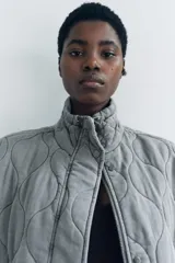 Chaleco gris acolchado con cuello alto, cierre frontal con cremallera metálica, bolsillos de plastrón en delantero y bajo ajustable con cordones.