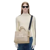 Cartera tote bag Marc Jacobs modelo The Medium Tote Bag, confeccionada en algodón color beige. Cuenta con asas superiores, correa cruzada ajustable y desmontable en color negro, y el texto impreso en contraste en el frente.