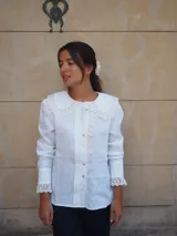 Camisa de lino color beige, con cuello bobo y detalles de encaje bordado en el cuello, pechera y puños. Cuenta con mangas largas levemente abuchonadas y cierre frontal con botones nacarados.