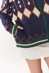 Campera tipo bomber tejida con diseño jacquard geométrico en tonos azul marino, blanco, rojo y verde. Presenta cierre frontal completo, cuello, puños y ruedo acanalados en color verde y blanco, con corte oversized y hombros caídos.