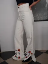 Pantalón vaquero de corte recto y tiro alto en color blanco marfil, con detalles de costuras en contraste y estampa decorativa en la parte inferior de las piernas, que incluye figuras de caballos y patrones geométricos. Presenta aberturas laterales en los tobillos.