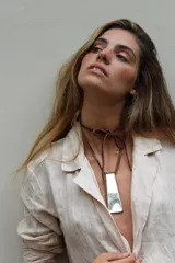 Gargantilla de diseño artesanal compuesta por un cordón fino de cuero marrón atado al cuello en forma de lazo y un dije colgante metálico de forma rectangular alargada y terminación irregular plateada.