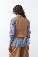 Chaleco de gamuza color marrón, con escote en V y cierre frontal con botones forrados. Tiene un largo a la cintura y silueta definida.