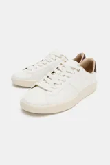 Zapatilla deportiva tipo bamba, color blanco, con suela plataforma engomada de color crema. Presenta pespuntes en el cuerpo y detalle en la talonera de color marrón oscuro.