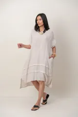 Vestido midi de lino color beige, de corte holgado y silueta acampanada. Cuenta con cuello redondo, mangas cortas amplias, bolsillos laterales y detalles de recortes horizontales en la falda con terminación asimétrica más larga en la parte trasera.