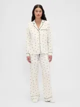 Conjunto de pijama compuesto por una camisa de manga larga con cuello solapa, cierre frontal de botones y bolsillo en el pecho, y un pantalón de corte recto con cintura elástica. Ambas piezas presentan un estampado de pequeños motivos oscuros sobre fondo claro y detalles de ribetes en contraste.