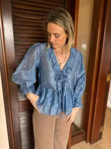 Blusa de tela satinada color azul, con escote en V, volados decorativos en el frente y mangas largas abullonadas con puños ajustados. Presenta un lazo frontal para atar y un diseño con volado en el ruedo.